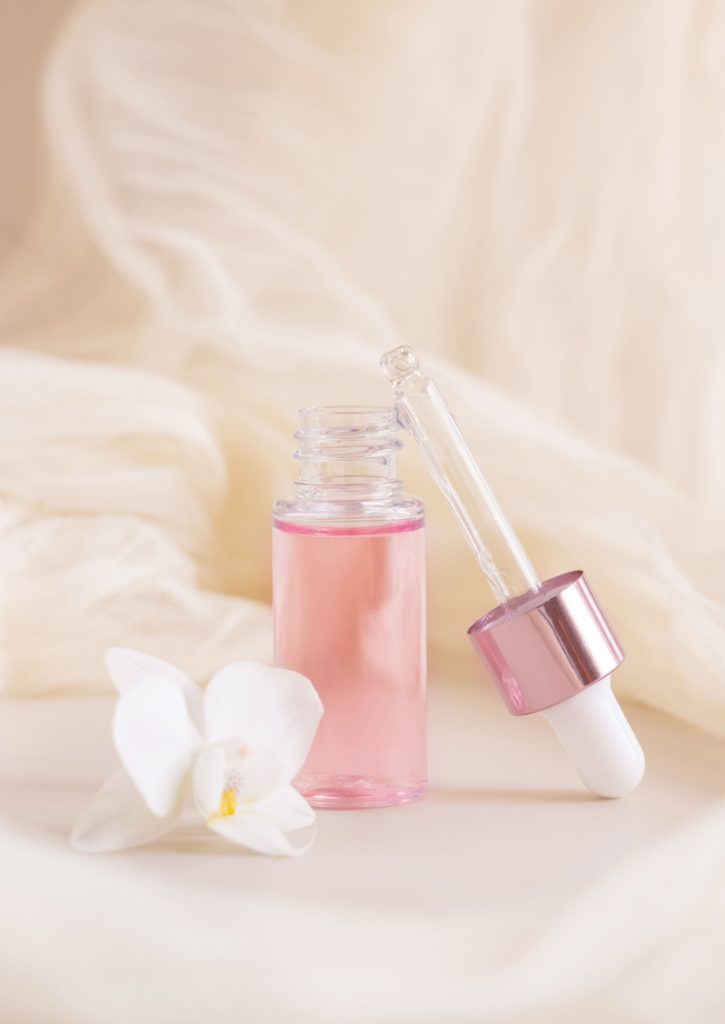 Pink Dropper Bottle near white orchid flower on light yellow close up. Skincare beauty product